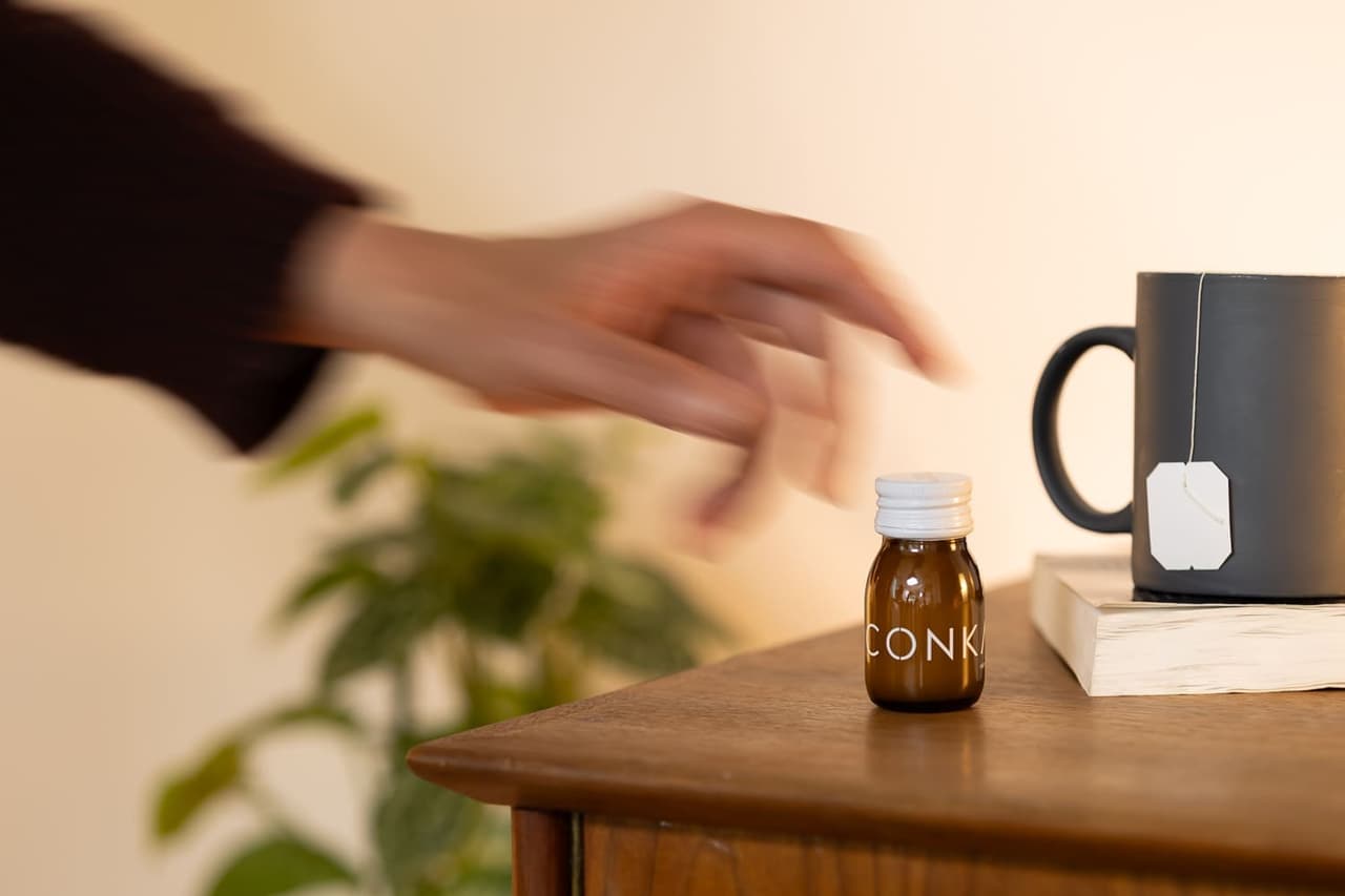 A hand reaching for a CONKA shot on a desk beside a mug and a book